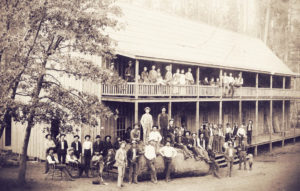 View of a two-story hotel with numerous people standing and sitting in front of it and on the balcony